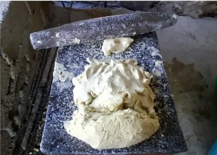 Fotografía de una tabla de amasar con masa de harina, un rodillo enharinado y un trozo de masa aparte, sobre una superficie de piedra en un espacio rústico.