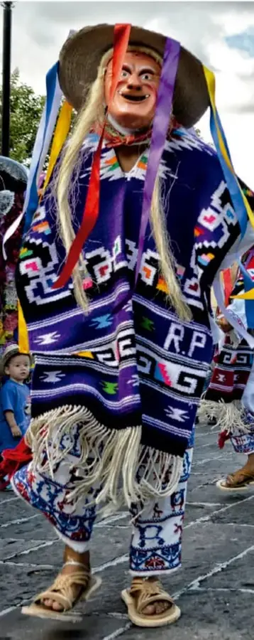 Persona disfrazada con máscara, sombrero de ala ancha con listones de colores, poncho tejido morado con iniciales 'R.P.' y huaraches; probablemente personaje de danza tradicional mexicana.