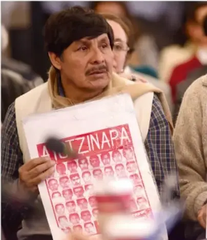 Persona sosteniendo un cartel con la palabra AYOTZINAPA y fotografías de personas desaparecidas, en un acto público de reclamo de derechos humanos.