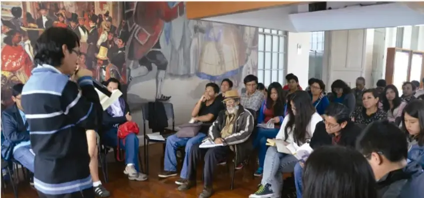 Fotografía de una asamblea escolar: un estudiante de pie con micrófono frente a un grupo de compañeros y adultos sentados en círculo, en un aula con mural histórico al fondo; varios participantes toman notas en sus cuadernos.