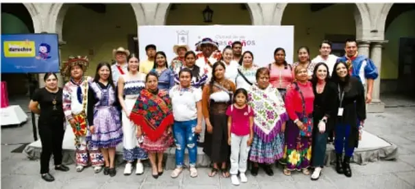 Fotografía grupal de comunidad escolar con niñas, niños y adultos, algunas personas con vestimenta tradicional, frente a un muro con logotipos institucionales.