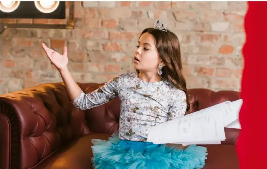 Fotografía de una niña con corona y vestido de fiesta sentada en un sillón, sosteniendo un guion impreso y con la mano en alto, como si estuviera ensayando una lectura dramatizada frente a una pared de ladrillo.