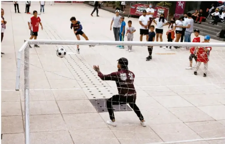 Fotografía de un grupo de niños y jóvenes jugando fútbol urbano en una plaza; un portero intenta detener el balón frente a la portería, con espectadores alrededor.