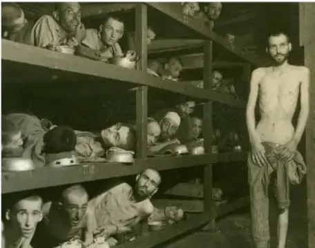 Fotografía en blanco y negro de prisioneros extremadamente delgados reclinados en literas de madera de tres niveles en un campo de concentración nazi, con un hombre de pie a la derecha mirando a la cámara.