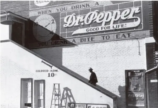 Fotografía en blanco y negro de un edificio con un anuncio publicitario de la bebida Dr. Pepper. En la parte inferior izquierda se lee 'COLORED-ADM 10c', letrero que indica una entrada segregada para personas afrodescendientes. Un hombre afrodescendiente camina frente al edificio.