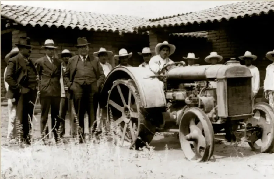 Fotografía histórica en sepia: grupo de campesinos y autoridades con sombreros junto a un tractor, frente a una construcción con techo de teja; entrega de tractores a campesinos, ca. 1940, Acervo INEHRM.