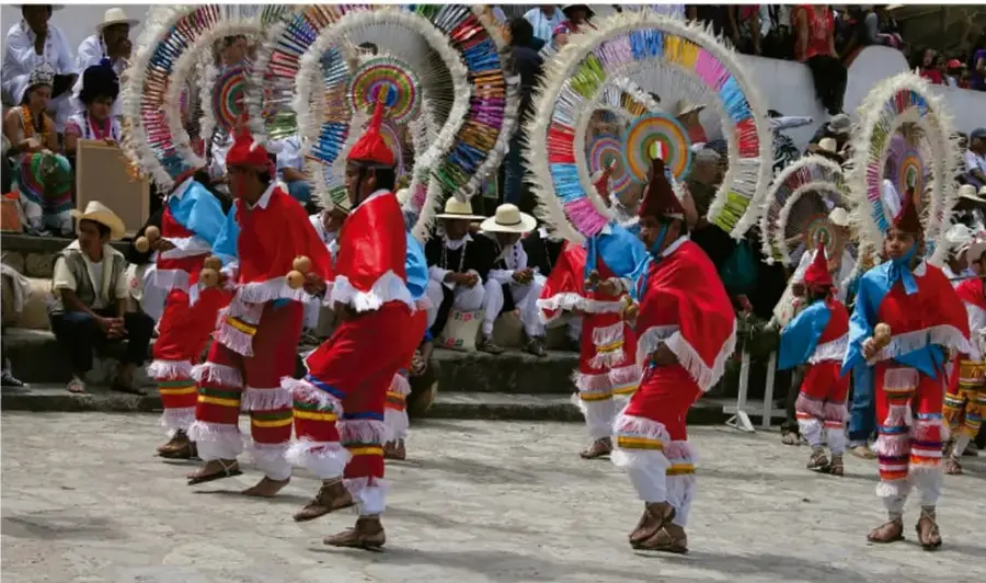 Fotografía de bailadores con trajes tradicionales rojos y blancos, con tocados circulares de plumas de colores (rojo, verde, azul, amarillo, rosa) y cascabeles en las piernas, danzando en una plaza mientras el público los observa desde gradas.