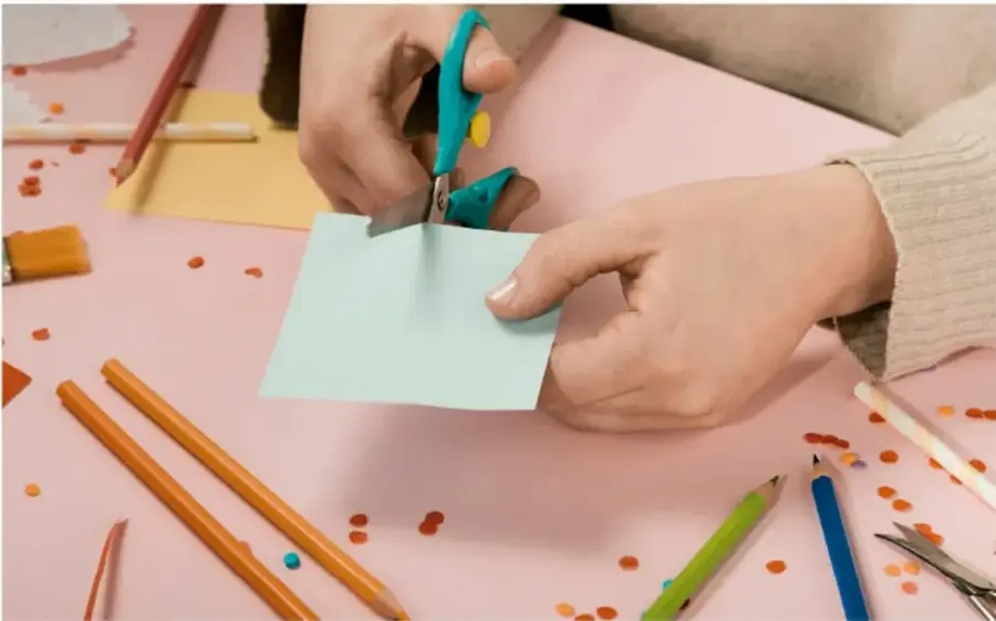 Fotografía de manos de un estudiante recortando un rectángulo de papel verde claro con tijeras azules sobre una mesa rosa, rodeado de lápices de colores, goma, sacapuntas y confeti.