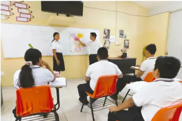 Fotografía de un aula de telesecundaria donde dos alumnos presentan frente a un grupo sentado en sillas naranjas; uno de ellos señala un cartel en el pizarrón.