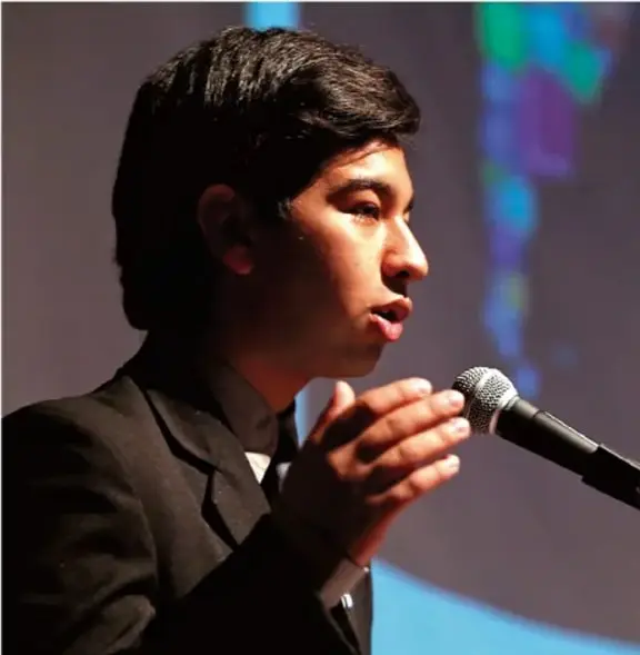 Fotografía de un adolescente hablando frente a un micrófono, con fondo azul y una pantalla con gráficos. Representa la participación juvenil en un evento público.