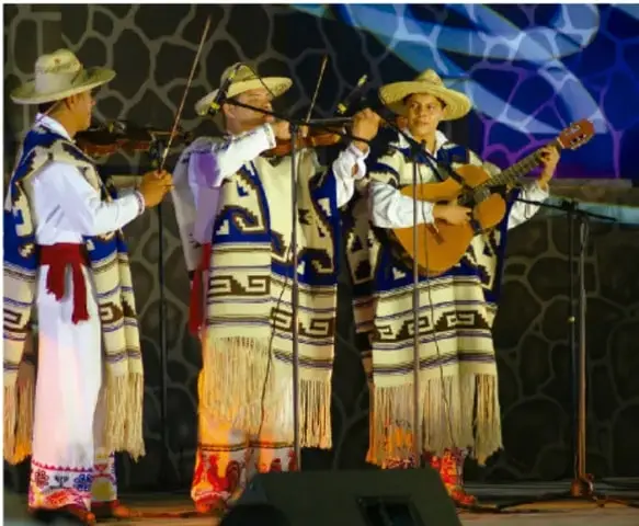 Fotografía de tres músicos p'urhépecha con vestuario tradicional (jorongos y sombreros), tocando violín y guitarra sobre un escenario con fondo azul, ilustrando la interpretación de pirekuas.