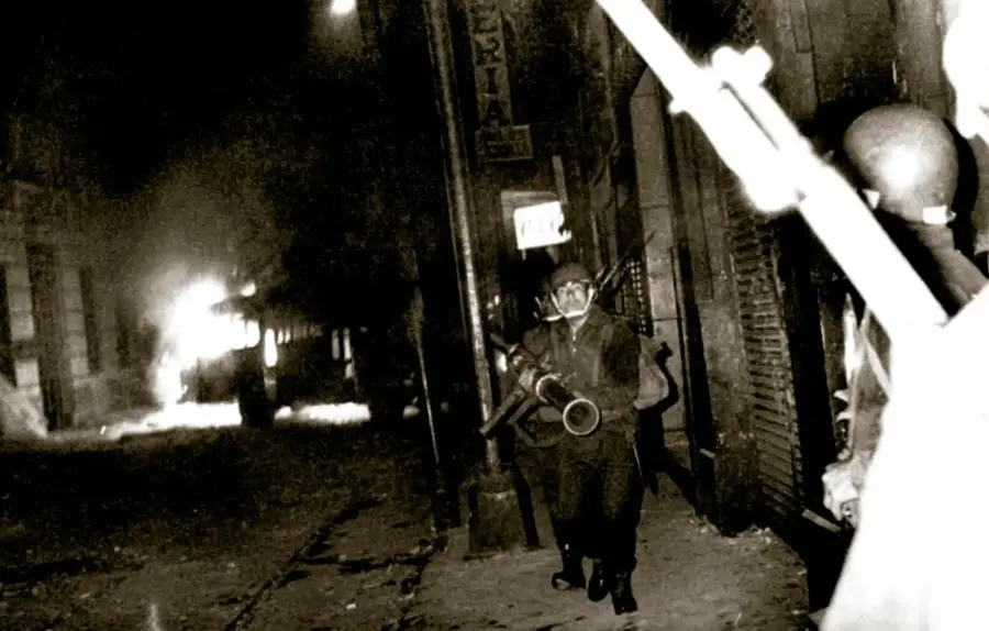 Fotografía nocturna en blanco y negro: soldados con casco, chaleco y armamento listos frente a la puerta de un edificio (Escuela Nacional Preparatoria), con llamas y humo al fondo en la calle; un segundo soldado aparece en primer plano a la derecha. Pie de foto: 'Soldados a punto de destruir la puerta de la Escuela Nacional Preparatoria, 30 de julio de 1968. Archivo Fotográfico de El Nacional, Fondo Temático, sobre: 0337-G (010). Acervo INEHRM.'