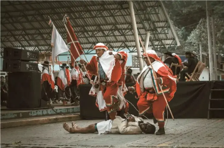 Fotografía de una ceremonia o danza tradicional indígena de Chiapas: varios participantes con trajes rojos y blancos y sombreros, uno tendido en el suelo, bajo una estructura techada con público alrededor.