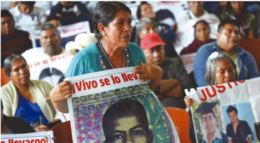 Fotografía de una asamblea de familiares de personas desaparecidas: una mujer sostiene un periódico o cartel con el texto "¡Vivo se lo llevaron!" y el retrato de un joven; al lado se ve otro cartel con la palabra "JUSTICIA" y fotografías de jóvenes, uno con birrete de graduación.