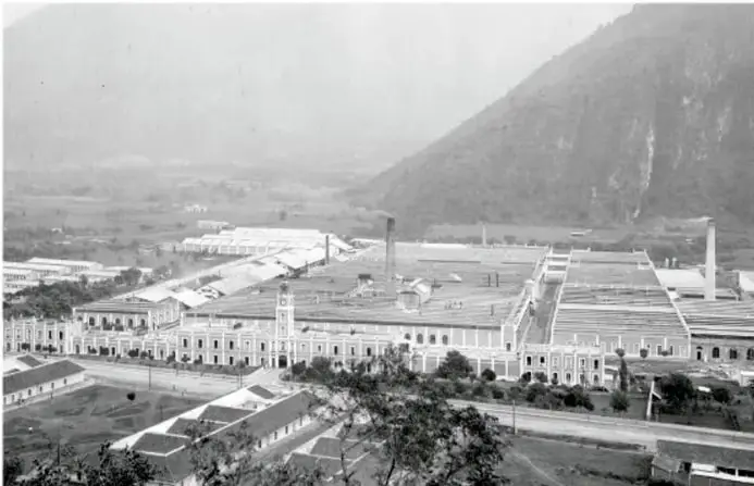 Fotografía histórica en blanco y negro de una gran fábrica o complejo industrial con montañas al fondo, probablemente durante el Porfiriato.