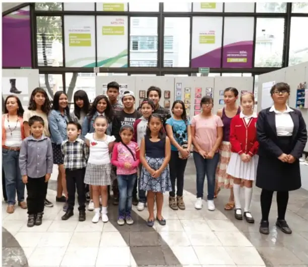 Fotografía de un grupo de estudiantes de secundaria, con uniforme, posando en un patio escolar con vitrinas de fondo.