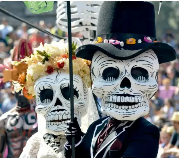 Dos calaveras (catrinas) con vestimenta de novia y sombrero, adornadas con flores, en una celebración pública del Día de Muertos.