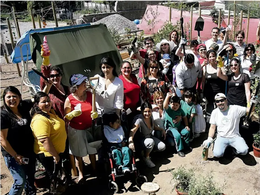 Fotografía de un grupo numeroso de personas de distintas edades (comunidad) con herramientas de jardinería (palas, guantes, plantas) en un terreno, aparentemente participando en una jornada comunitaria de siembra o huerta.