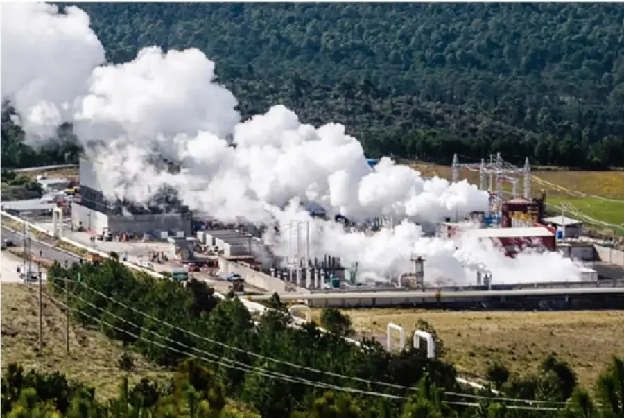 Fotografía aérea de una central geotérmica rodeada de vegetación, con grandes columnas de vapor blanco provenientes de las torres de enfriamiento y líneas eléctricas cercanas.