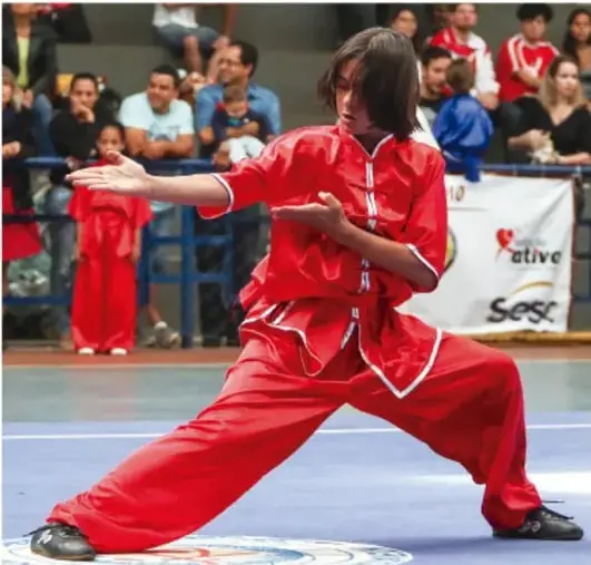 Estudiante con uniforme rojo de artes marciales practicando wushu o kung fu en un evento deportivo con público al fondo.