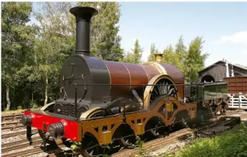 Fotografía de una locomotora de vapor antigua (motor de la Segunda Revolución Industrial) sobre rieles, con fondo de árboles.
