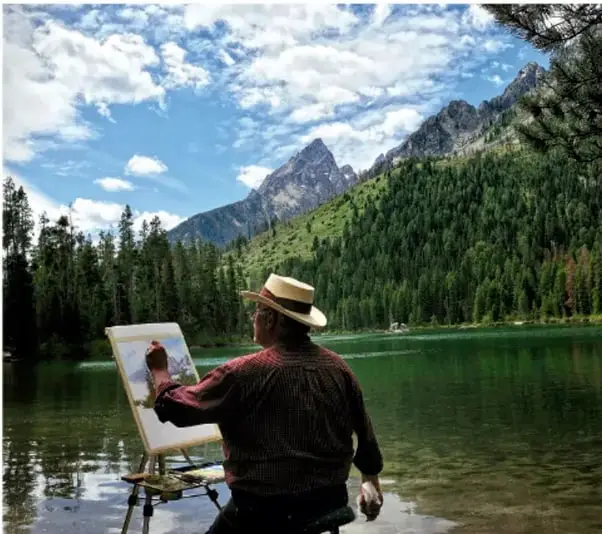 Fotografía de un artista con sombrero de paja pintando un paisaje con caballete frente a un lago rodeado de montañas y bosque, bajo un cielo nublado.