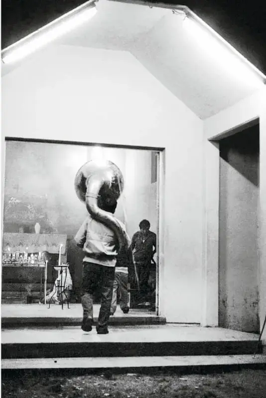 Fotografía en blanco y negro de una persona de espaldas entrando a un taller o estudio, portando un objeto voluminoso sobre el hombro, con otra persona observando al fondo y una mesa con objetos.