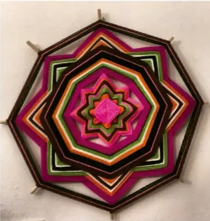 Textil circular en forma de estrella u octógono de capas concéntricas, con hilos de colores rosa, fucsia, verde, naranja y negro, sostenido por varillas en los vértices; parece un ejemplo de arte textil o telar circular comunitario.
