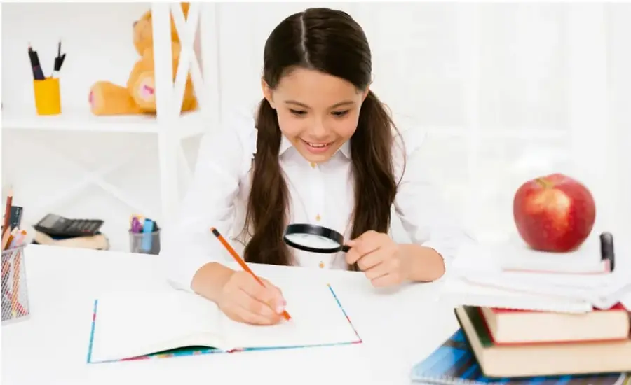 Fotografía de una estudiante de secundaria sentada en su escritorio, sonriente, sosteniendo un lápiz y una lupa mientras escribe en un cuaderno; sobre la mesa hay libros apilados, un manzana roja, lápices en un portapapeles y un oso de peluche en un estante al fondo.