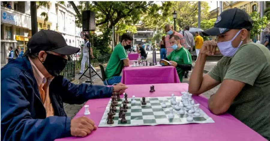 Fotografía de dos personas jugando ajedrez al aire libre en una plaza, con cubrebocas, sobre mesas con mantel rosa; al fondo hay más personas y edificios.