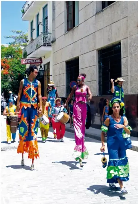 Comparsa o cortejo callejero con personas con vestimenta colorida y tambores, en una calle con edificios.