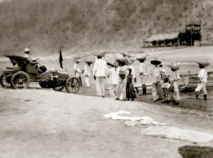 Fotografía histórica en sepia titulada 'Peones de Iguala, Guerrero, ca. 1910' del INAH/SINAFO. Muestra a un grupo de peones con sombreros y ropa blanca junto a un automóvil temprano, en un camino rural con montañas y una estructura de madera al fondo.