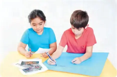 Dos estudiantes sentados a una mesa con papeles de color, revistas y tijeras, elaborando un papelógrafo.