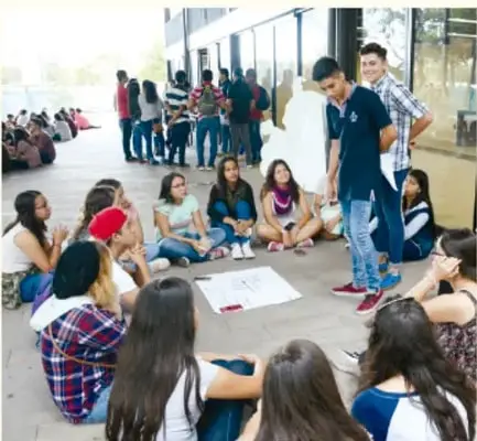 Estudiantes sentados en círculo en el patio escolar, dialogando y tomando notas sobre una hoja; un docente de pie observa.