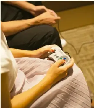 Fotografía de una persona sentada usando un control de videojuego, con otra persona al fondo.