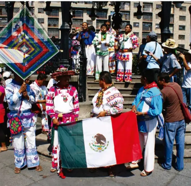 Fotografía de una comunidad indígena o pueblos originarios en una plaza urbana, portando una bandera de México y artesanías textiles multicolores, representando la identidad y la cultura.