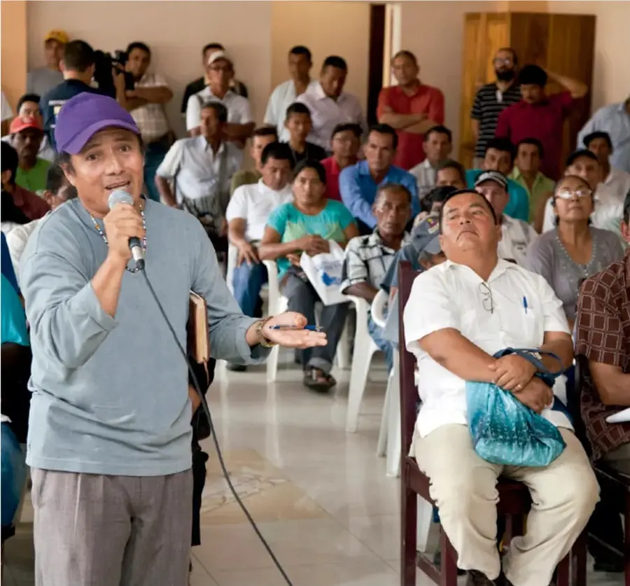 Fotografía de una asamblea comunitaria: una mujer con gorra violeta sostiene un micrófono y habla de pie, mientras un grupo diverso de personas la escucha sentado en sillas.