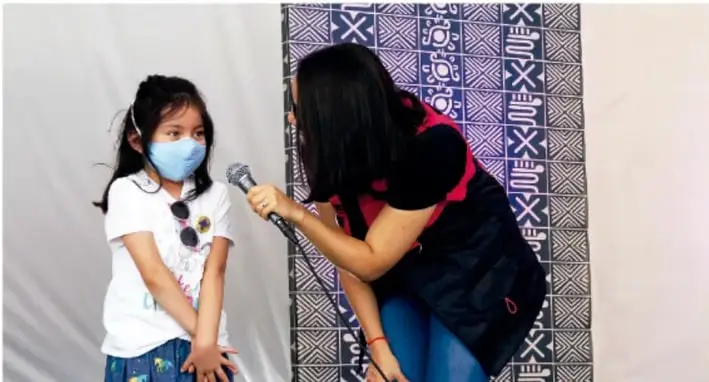 Fotografía de una niña con cubrebocas siendo entrevistada por una mujer joven que sostiene un micrófono; al fondo hay un muro con un tapete de diseños geométricos.