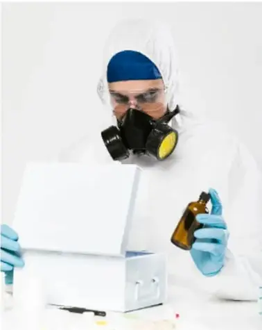 Persona con equipo de protección completa (mascarilla con filtro, traje blanco, guantes y gorro) manipulando un frasco ámbar.