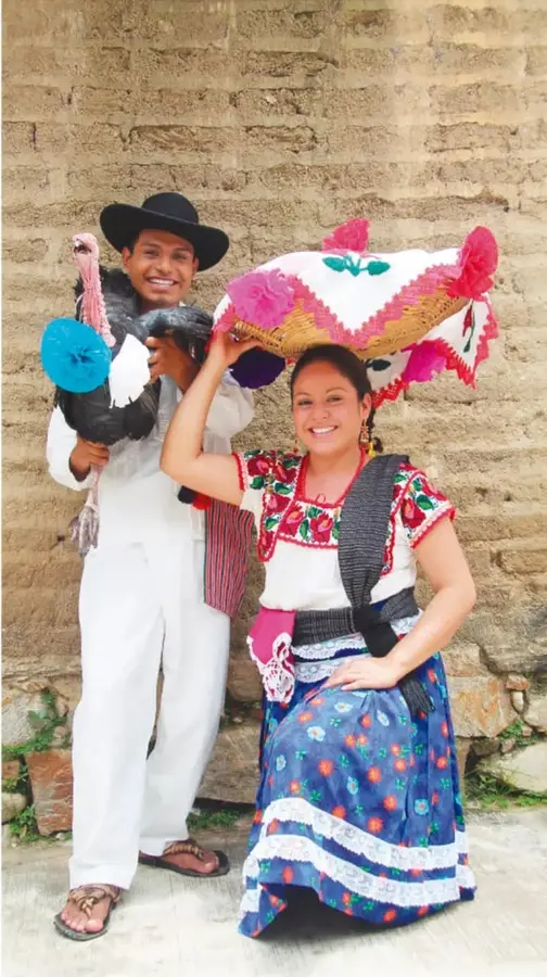Pareja sonriente con trajes tradicionales frente a un muro de piedra: el hombre de blanco y sombrero carga un guajolote vivo; la mujer con blusa bordada y falda azul lleva una canasta decorada con papel rosa sobre la cabeza.