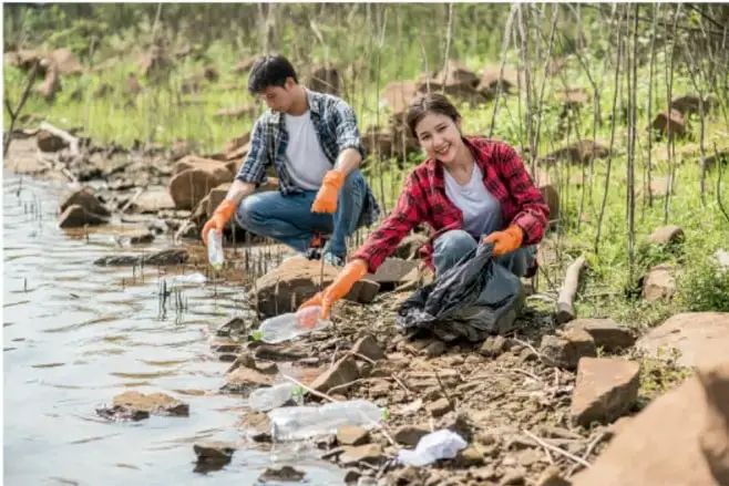 Fotografía de dos estudiantes con guantes recogiendo residuos (plásticos y basura) en la orilla rocosa de un río, rodeados de vegetación; ilustra el trabajo de campo del proyecto.
