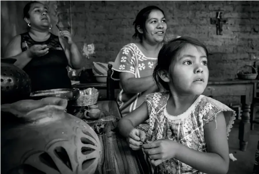 Fotografía en blanco y negro de una familia indígena mexicana en un taller de cerámica: una niña en primer plano observa mientras dos mujeres trabajan el barro.