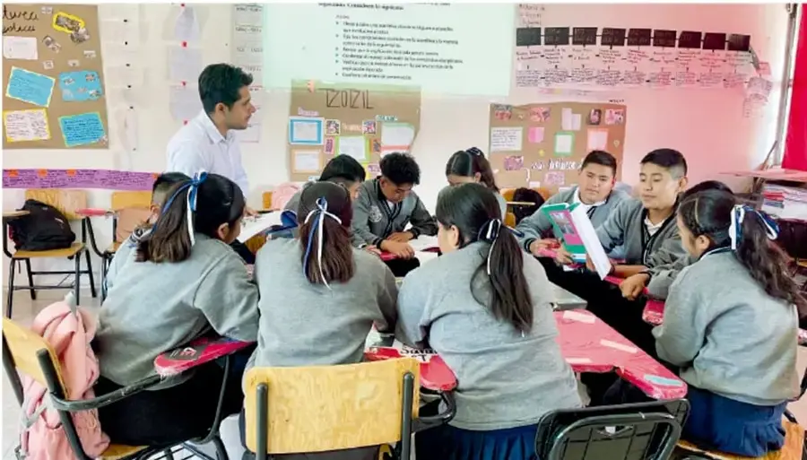 Fotografía de un grupo de estudiantes de telesecundaria reunidos en círculo alrededor de pupitres, trabajando en equipo en el aula; un docente los observa de pie junto a un pizarrón con carteles.