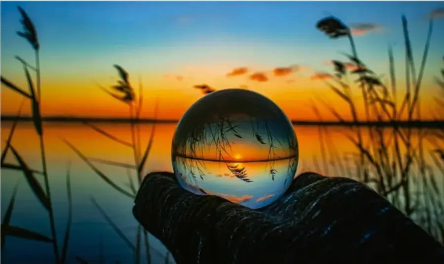 Fotografía de una mano enguantada sosteniendo una esfera de vidrio transparente que refleja un atardecer con juncos, sobre un fondo de cielo y agua al atardecer.