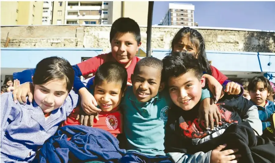 Fotografía de un grupo de niños y niñas de primaria abrazados y sonrientes en el patio de la escuela, con edificios de departamentos al fondo y cielo despejado.