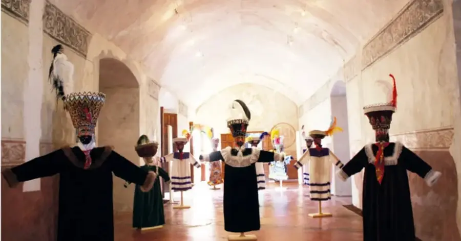 Fotografía del interior de un exconvento donde figuras disftradas con trajes y máscaras tradicionales (chinelos) están expuestas en una sala de museo.