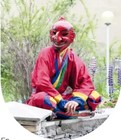 Persona sentada con la piernas cruzadas que viste una máscara roja de demonio o diablito con dientes blancos y traje colorido; al fondo, plantas y una farola.