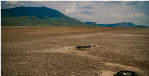 Fotografía de un paisaje árido con un lago o laguna seca, montañas al fondo y cielo nublado. En primer plano se observa un terreno seco y agrietado con algunos objetos oscuros.