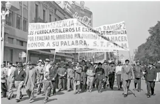 Fotografía en blanco y negro de una manifestación de ferrocarrileros. Los trabajadores marchan portando mantas con consignas como 'Los ferrocarrileros le exigimos la renuncia por lealtad al gobierno por dignidad y honor a su palabra. S.T.F.R.M.'