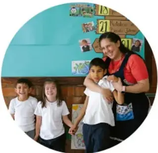 Fotografía circular de una maestra que abraza con cariño a un niño en el salón de clases, frente a un mural con fotografías, mientras dos alumnas de uniforme blanco los observan sonrientes.